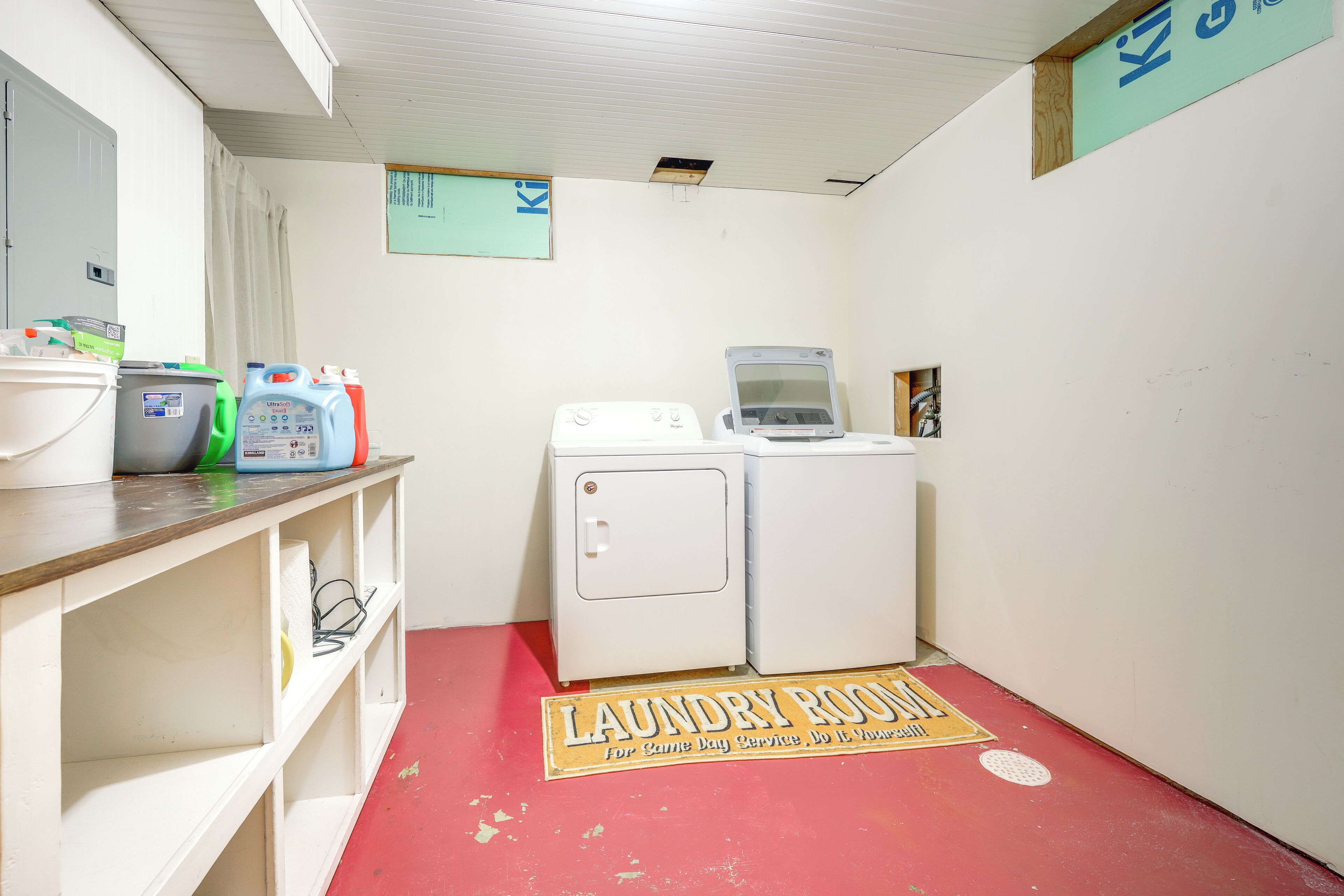A laundry room with a red floor, white washing machines, and shelves with cleaning supplies.