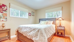 Un dormitorio con una cama de madera, dos ventanas, una lámpara de noche y una obra de arte floral en la pared.