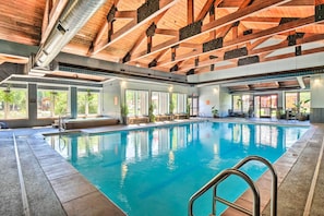 Una piscina interior con agua azul cristalina, vigas de madera en el techo y grandes ventanales que dejan entrar luz natural.