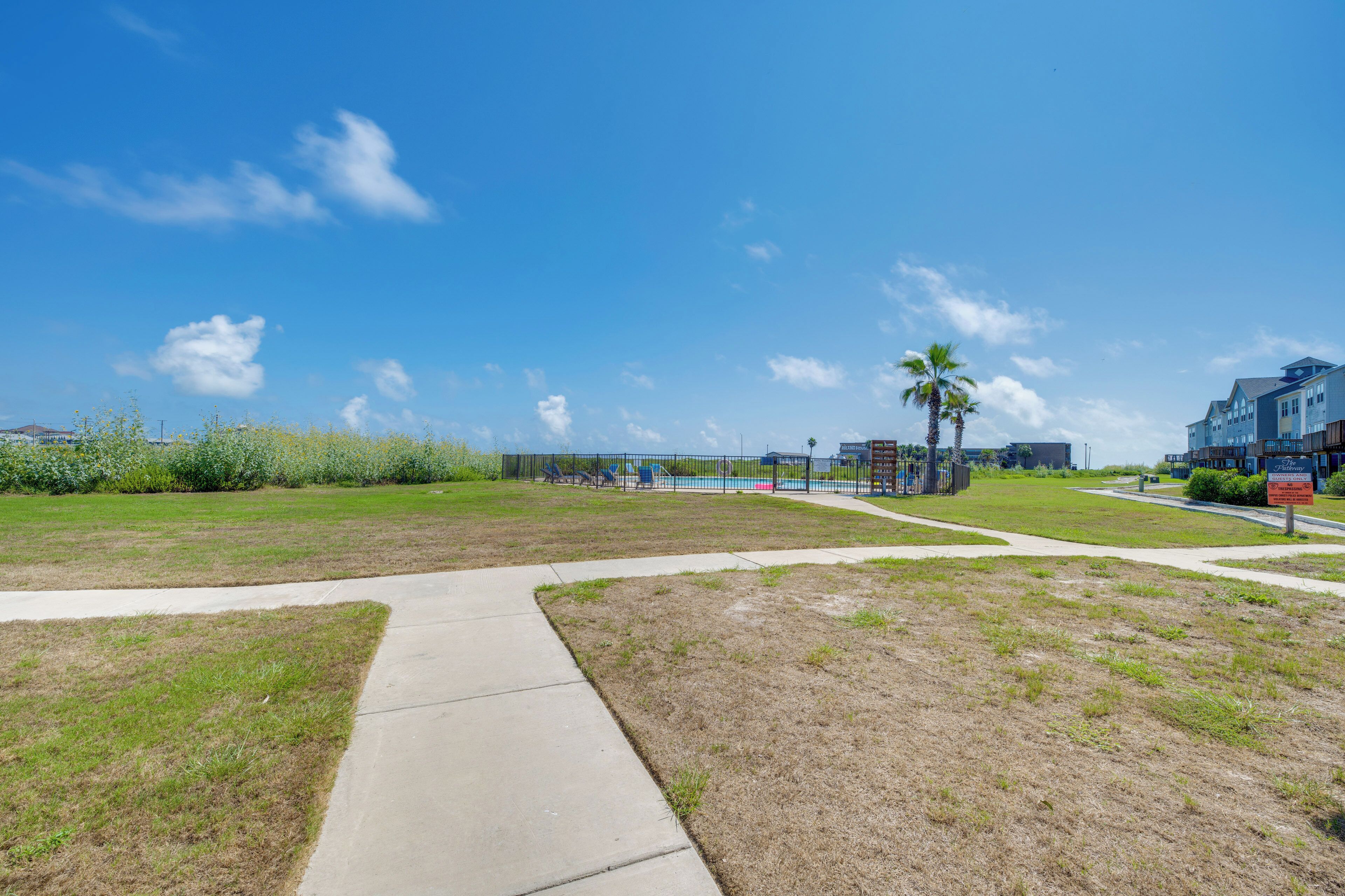 A grassy area with a concrete path leading to a pool, surrounded by buildings and palm trees.
