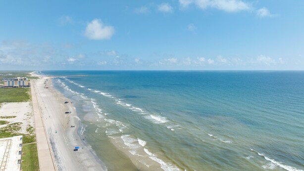 Ein Strand mit weitem Sandstreifen, einigen Sonnenschirmen und ein paar Personen. Im Hintergrund sind Gebäude zu sehen.