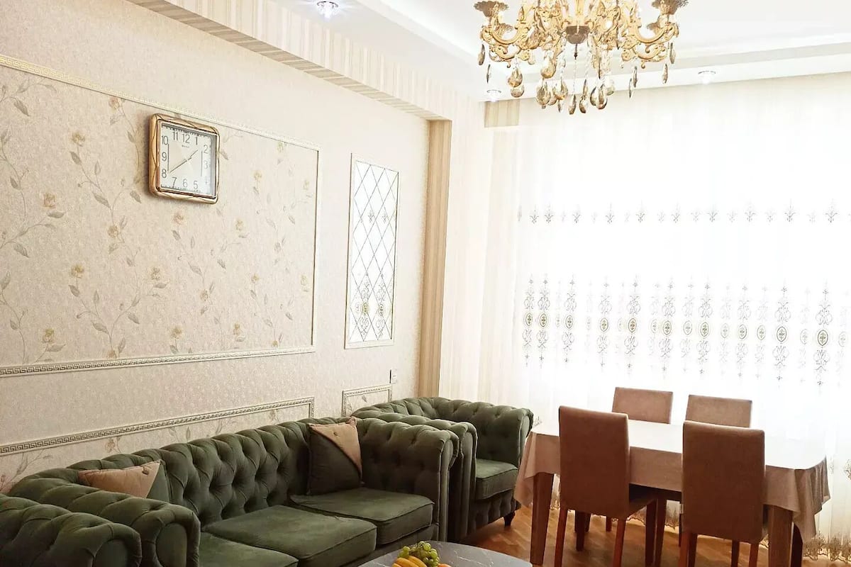 Ein Wohnzimmer mit einem grünen Sofagarnitur, einem Holztisch mit Früchten, einer Kristallleuchtre und einem Essbereich mit Tisch und Stühlen.