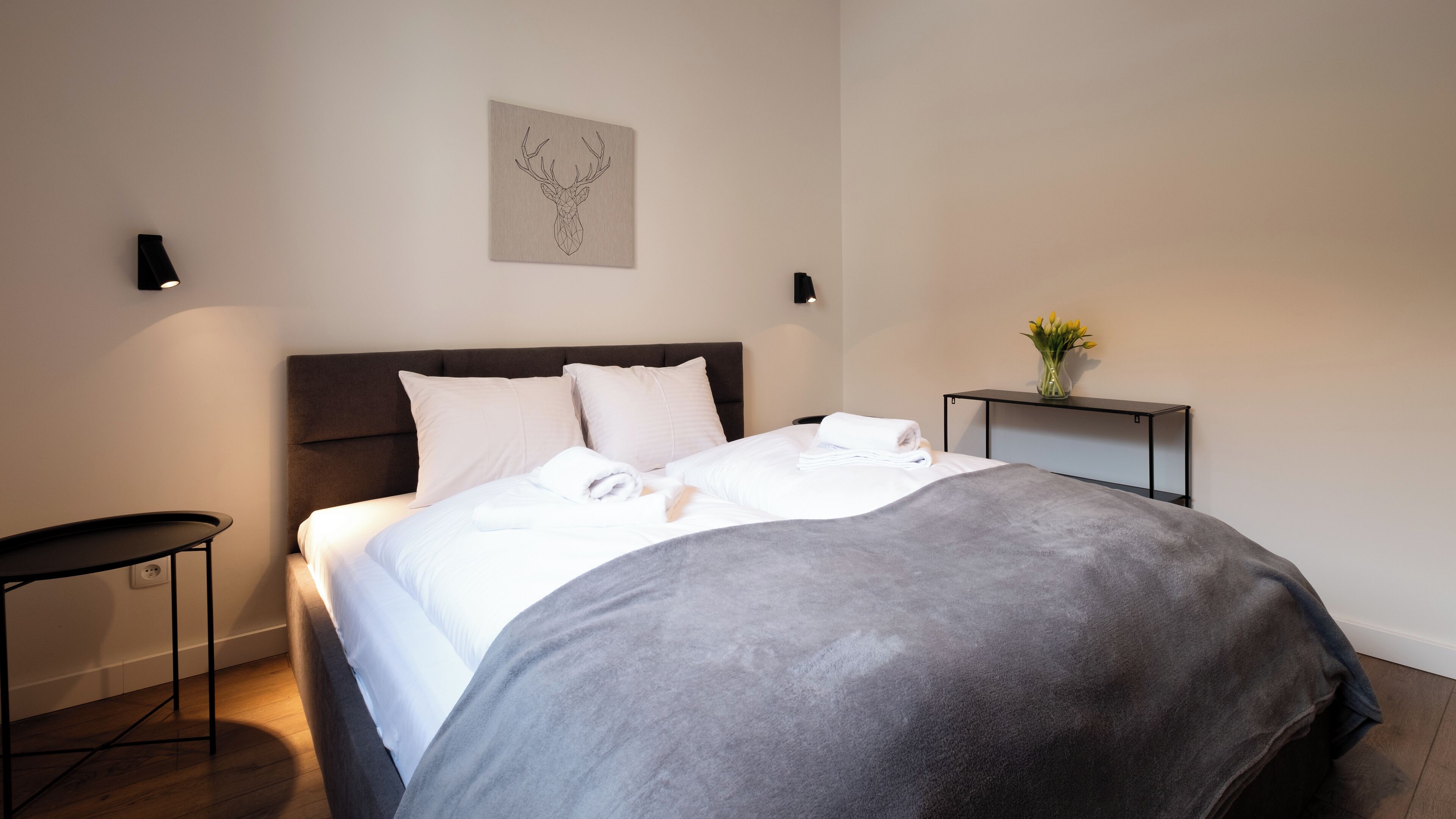A neatly made bed with a grey blanket, a bedside table with a vase of flowers, and a wall-mounted artwork.