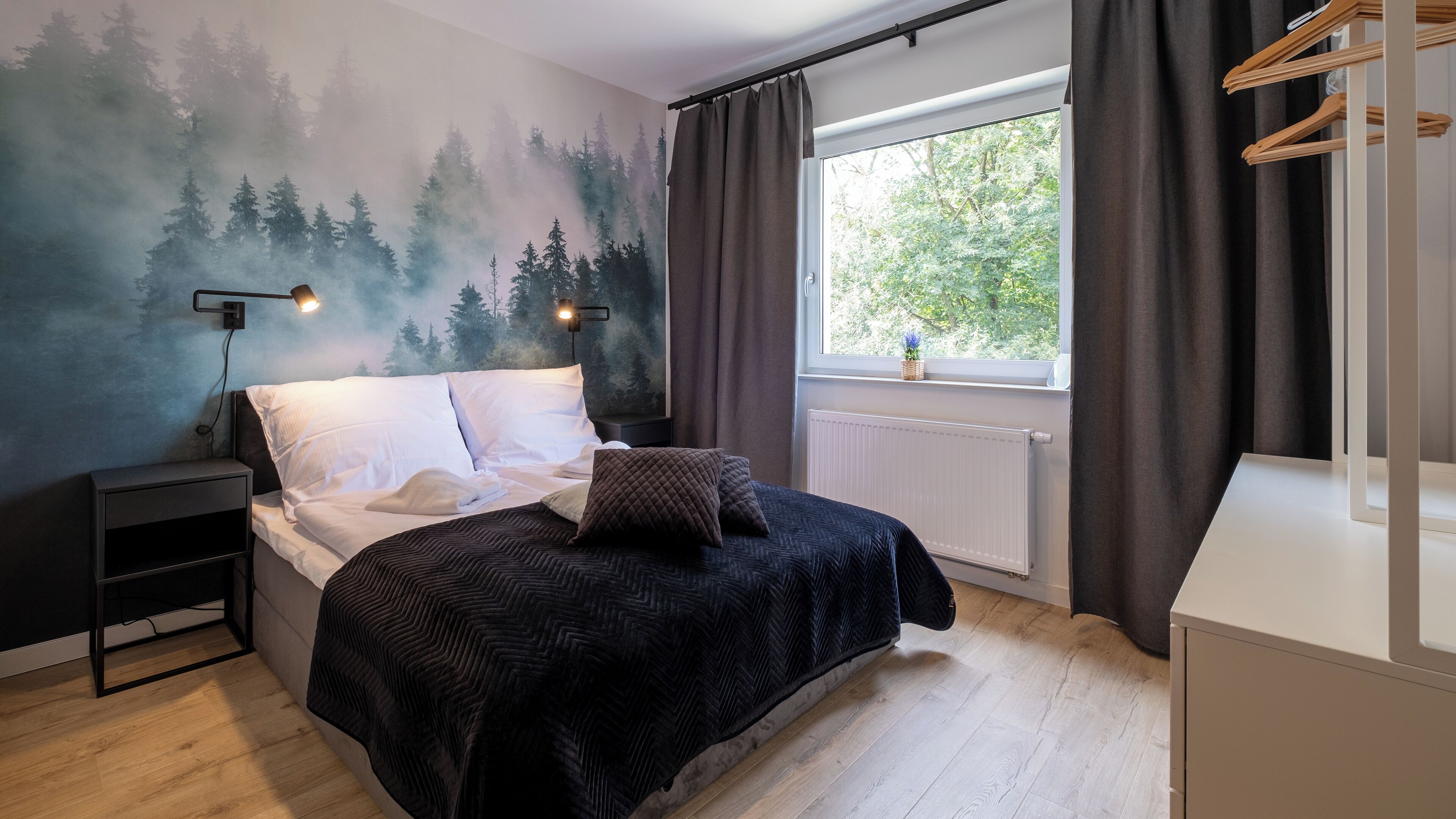A bedroom with a bed, a nightstand, a wall-mounted lamp, a window with curtains, and a view of trees outside.