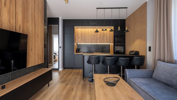 A modern kitchen with wooden cabinets, a central island with bar stools, a built-in oven, and a large flat-screen TV. - Apartamenty Sun & Snow River Park (Wisla)