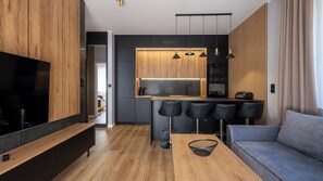 A modern kitchen with wooden cabinets, a central island with bar stools, a built-in oven, and a large flat-screen TV. - Apartamenty Sun & Snow River Park (Wisla)