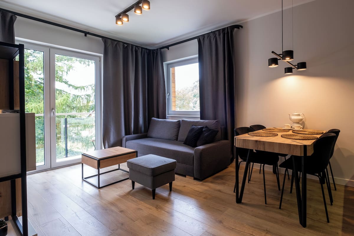 A modern living room with a grey sofa, a wooden dining table with black chairs, and a window with curtains.