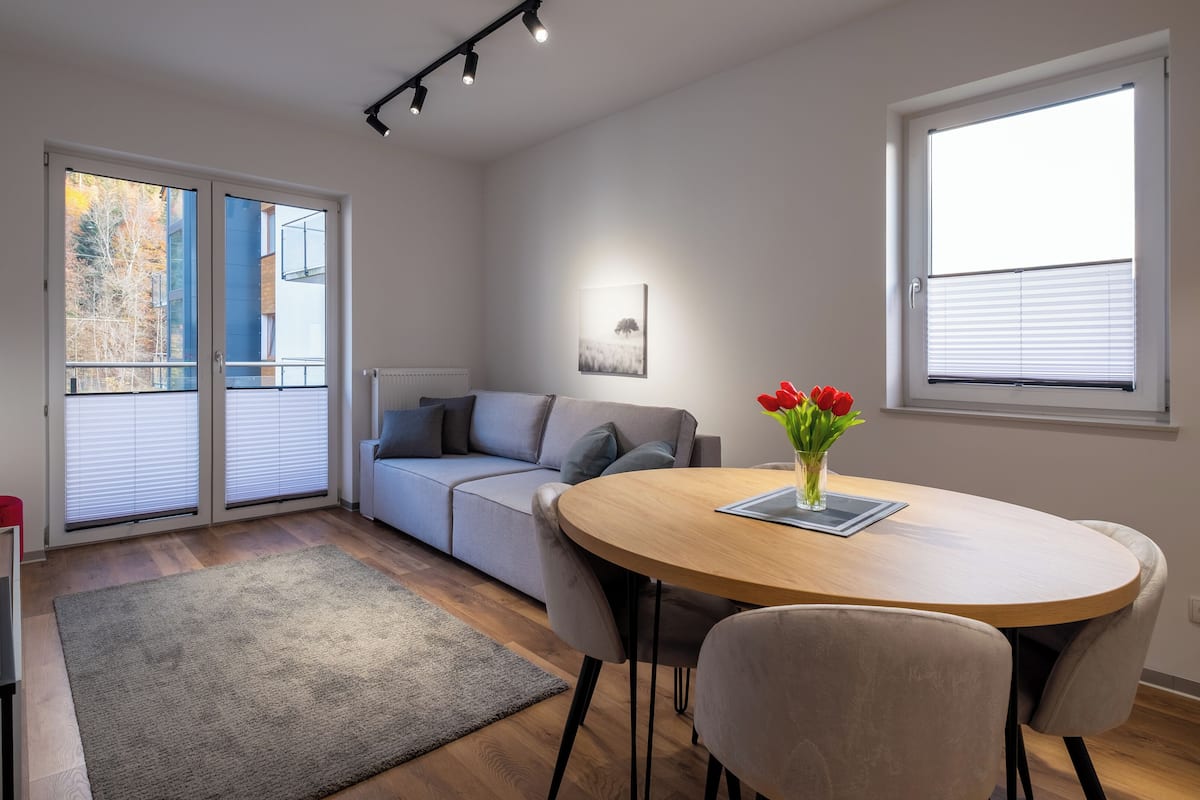 A modern living room with a sofa, a dining table with chairs, a glass coffee table, a TV, and a window with blinds.
