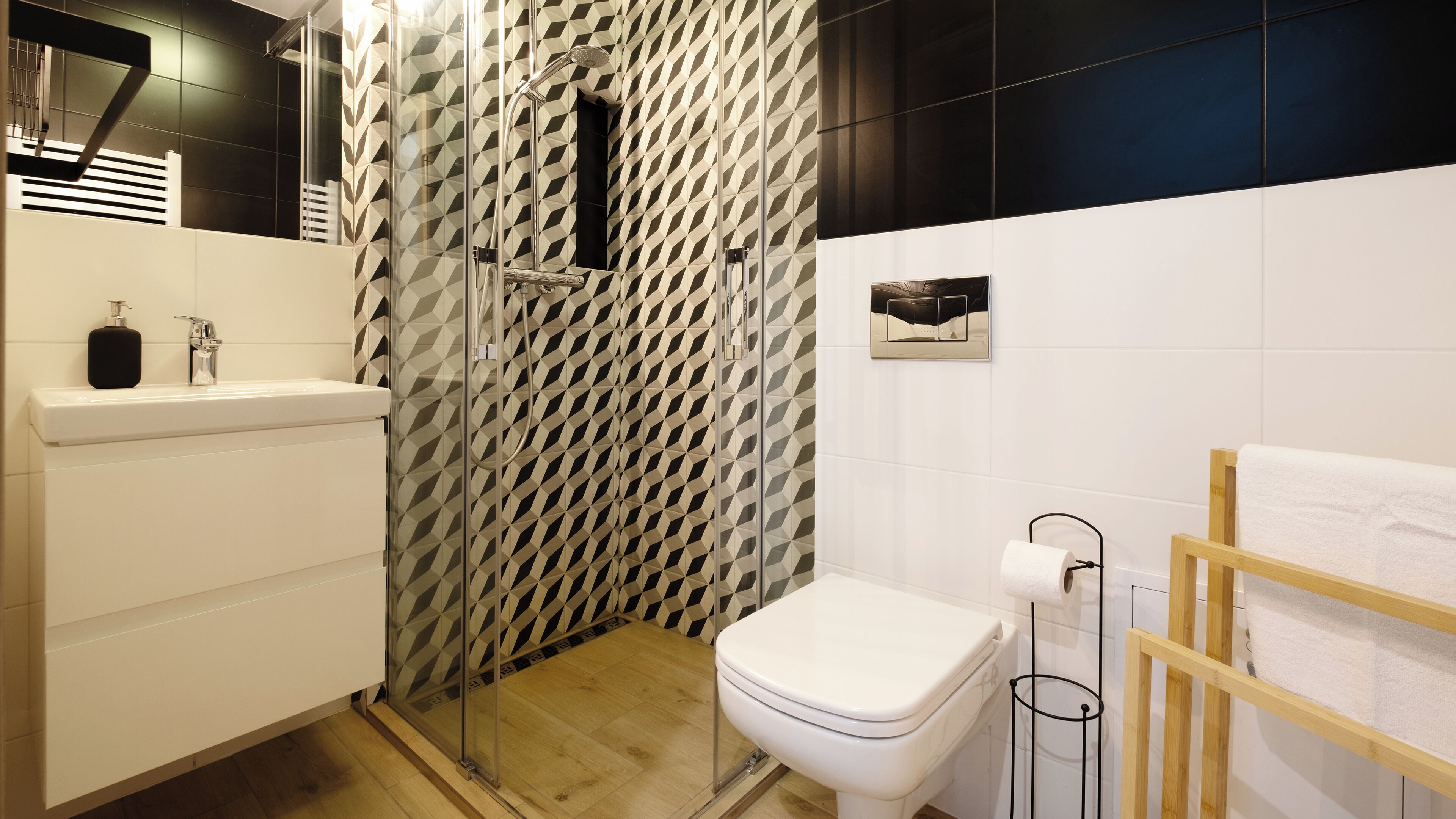 A modern bathroom with a geometric patterned shower wall, a white toilet, a wooden towel rack, and a white sink with a soap dispenser.