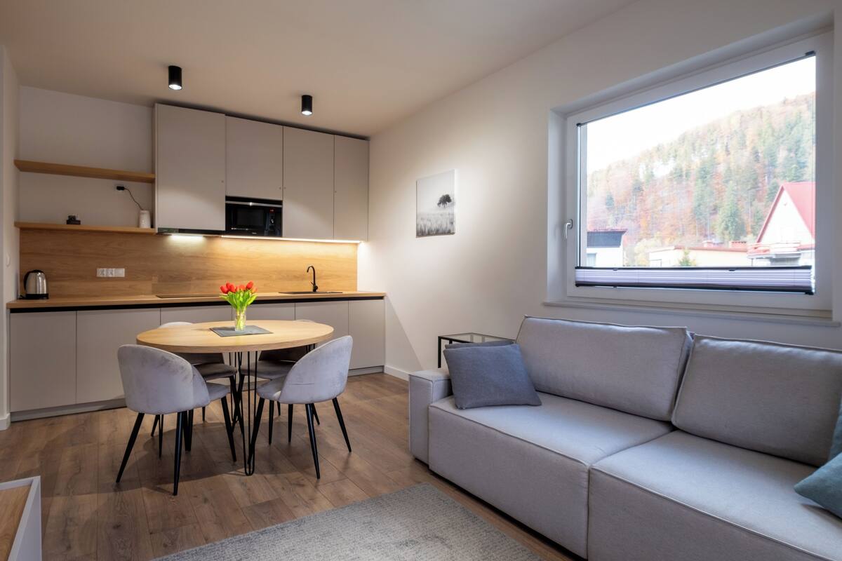 A modern kitchen with a dining area, a sofa, and a window with a view of trees.