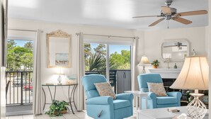 Une pièce avec deux fauteuils bleus, une table blanche, un ventilateur de plafond, une grande fenêtre donnant sur des arbres et une petite table avec une lampe.