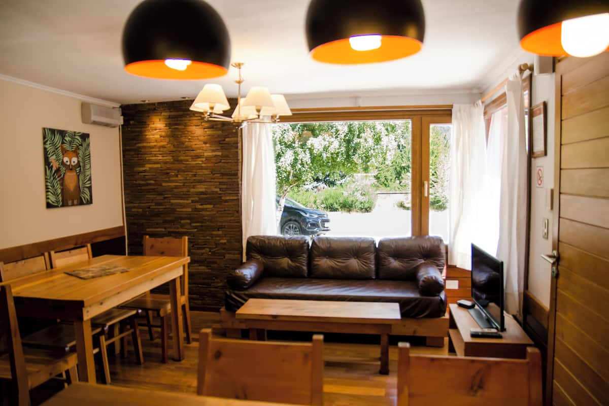 Ein gemütliches Zimmer mit einem Holzespeisetisch, einer Ledersofa und einem Fernseher. Eine Steinwand und ein Fenster mit Blick nach draußen sind ebenfalls vorhanden.