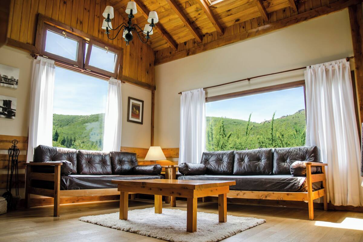 Ein Wohnzimmer mit Holzboden, einer Ledersofa, einem Holztisch und großen Fenstern mit Blick auf grüne Hügel.