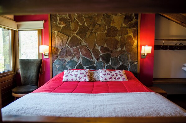 Una habitación con pared de piedra, colcha roja y una silla.