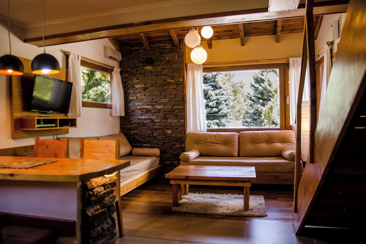 Ein gemütliches Wohnzimmer mit Steinwand, Holzbalken, einer Couch, einem Couchtisch und einem Fernseher auf einem Regal.