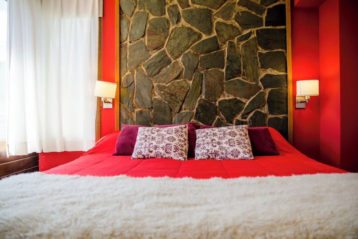 Ein Bett mit roter Bettdecke, weißen, flauschigen Kissen und einer Steinwand im Hintergrund.
