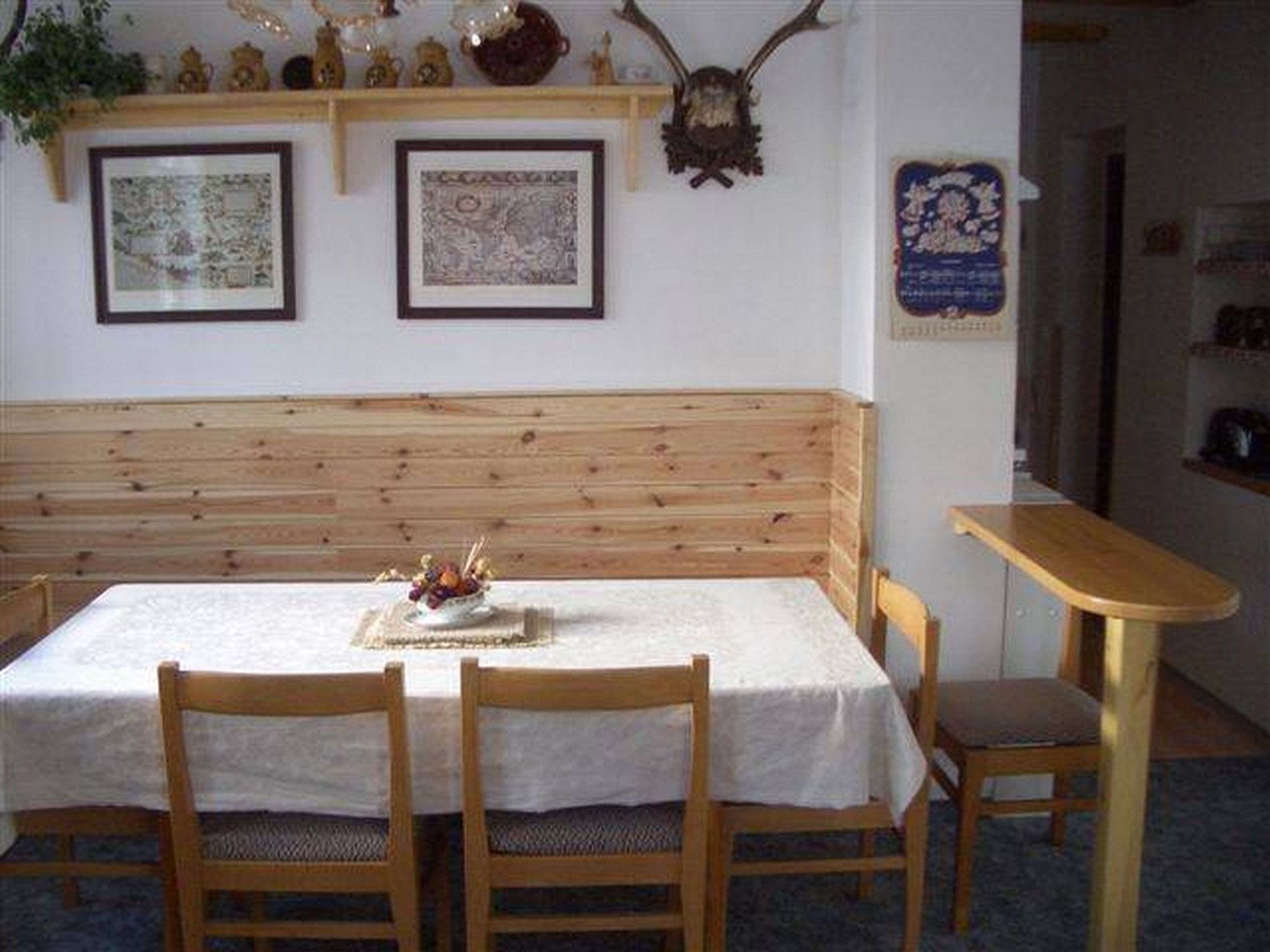 Een kamer met een houten bank, een tafel met een witte tafelkleed en twee stoelen. Er hangen ingelijste foto’s aan de muur en een decoratief object boven de bank.