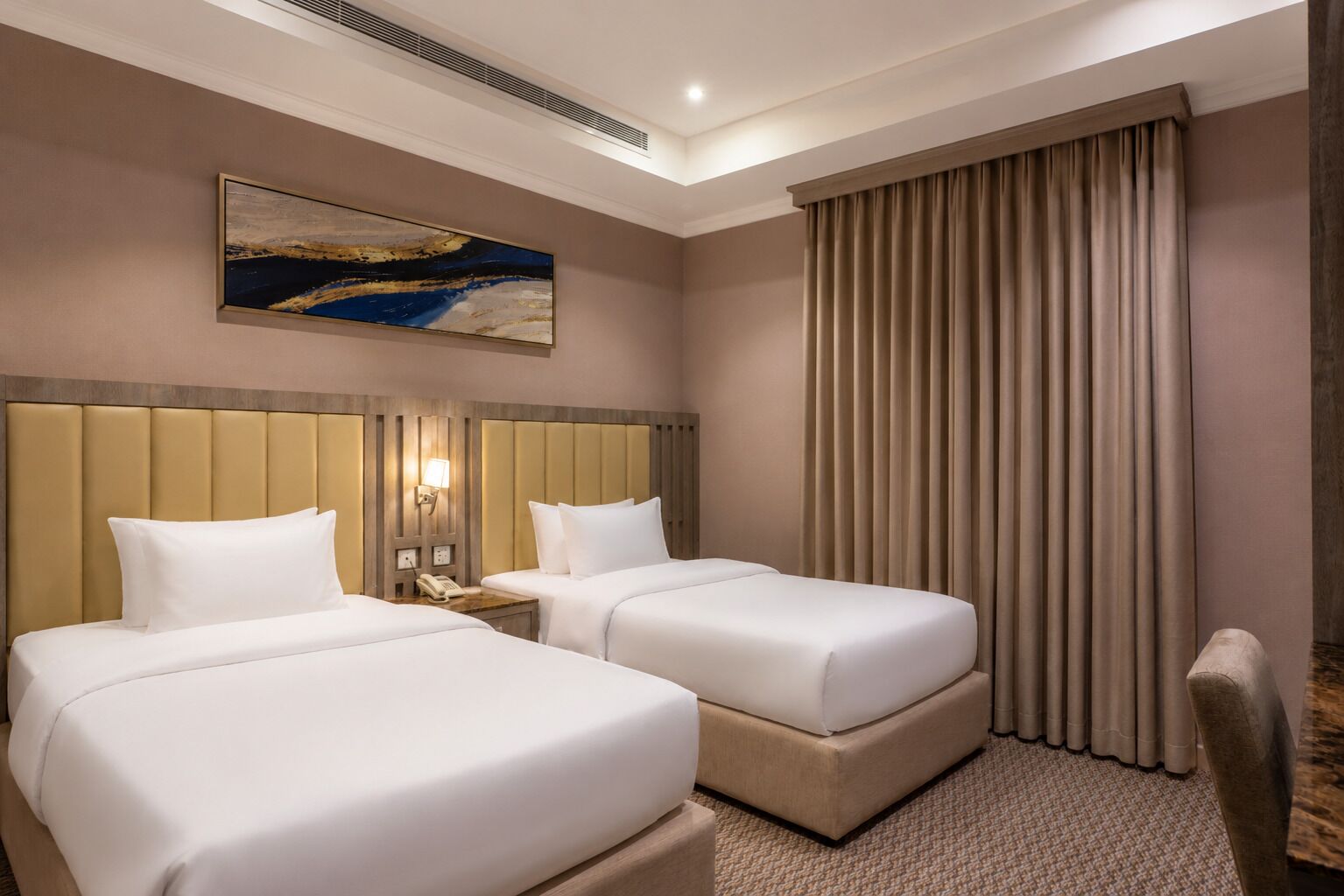 Habitación de hotel con dos camas, cabecera con líneas verticales, mesita de noche con lámpara, un cuadro en la pared y un ventanal con cortinas.