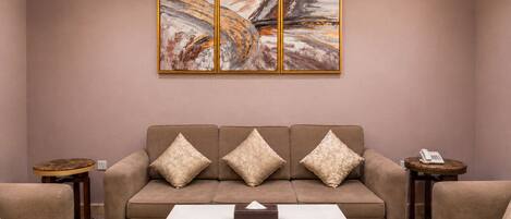 Una sala de estar con un sofá beige, tres cojines a juego, una mesa de centro blanca y un cuadro abstracto colgado en la pared.