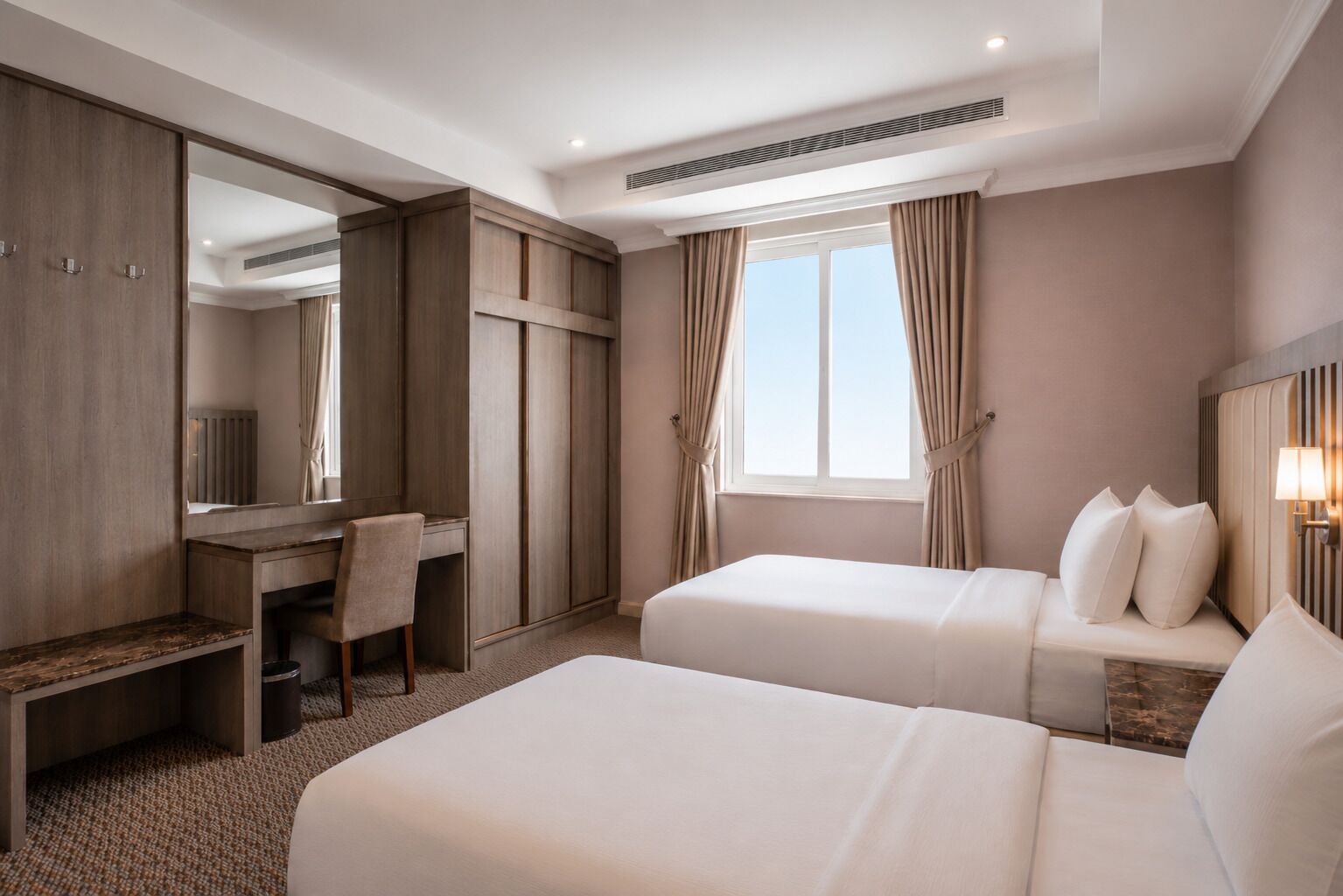 Ett hotellrum med två sängar, ett skrivbord, en stol, en stor spegel och ett fönster med gardiner.