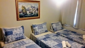 A room with three beds, each with patterned blue and white bedding. A framed painting of a sailing ship is on the wall. - KUMBH GANGA RETREAT (Karchana)