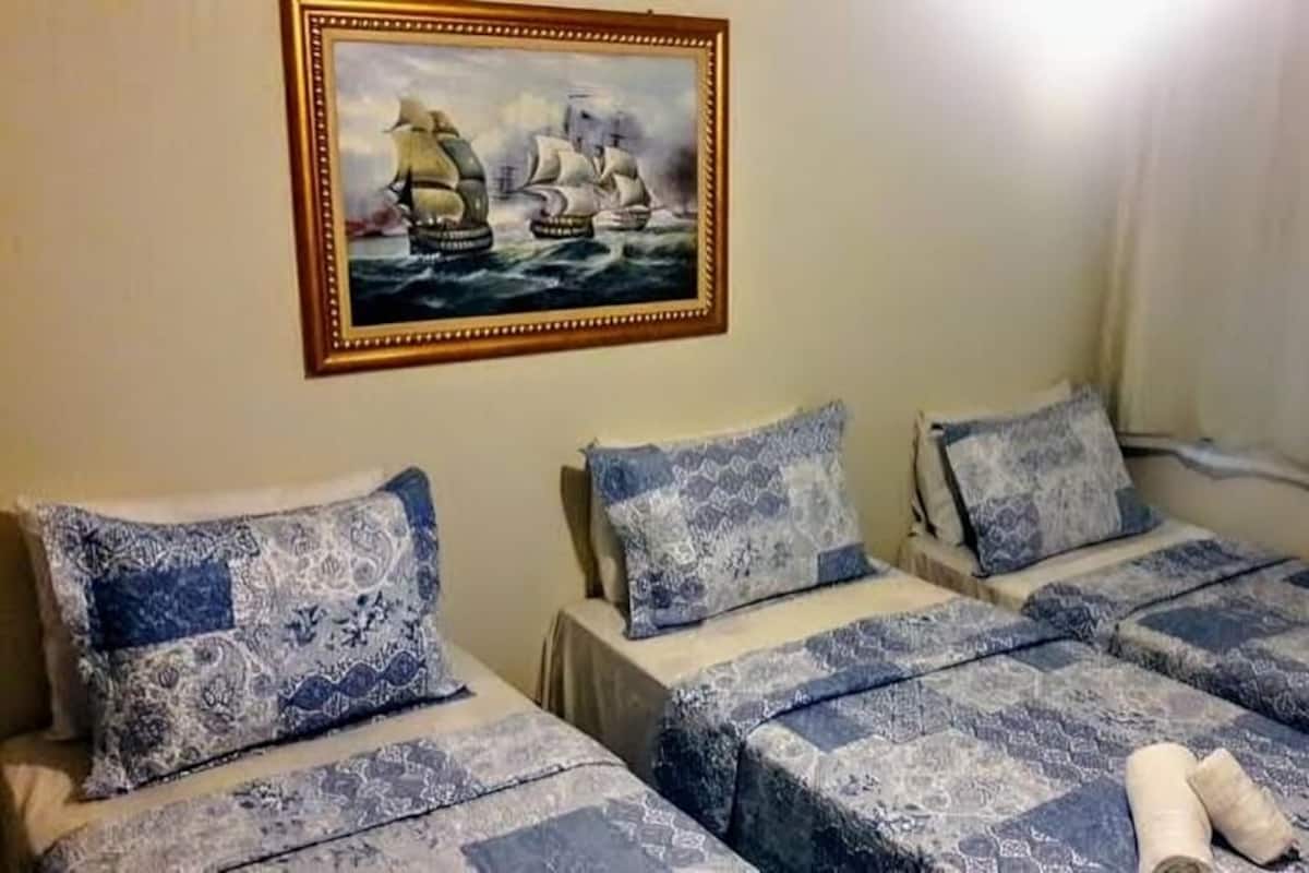 Quarto com três camas, cada uma com roupa de cama estampada em azul e branco. Um quadro de um navio à vela está pendurado na parede.