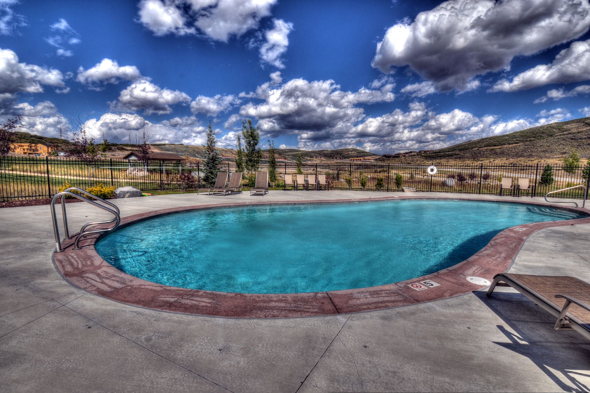 Una piscina con borde rojo, rodeada por una cerca, y un cielo azul despejado con nubes.