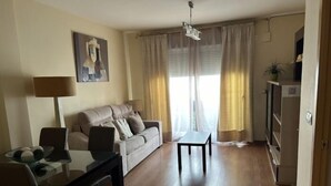 A living room with a sofa, a dining table, and a lamp. There is a painting on the wall and a plant on a shelf.