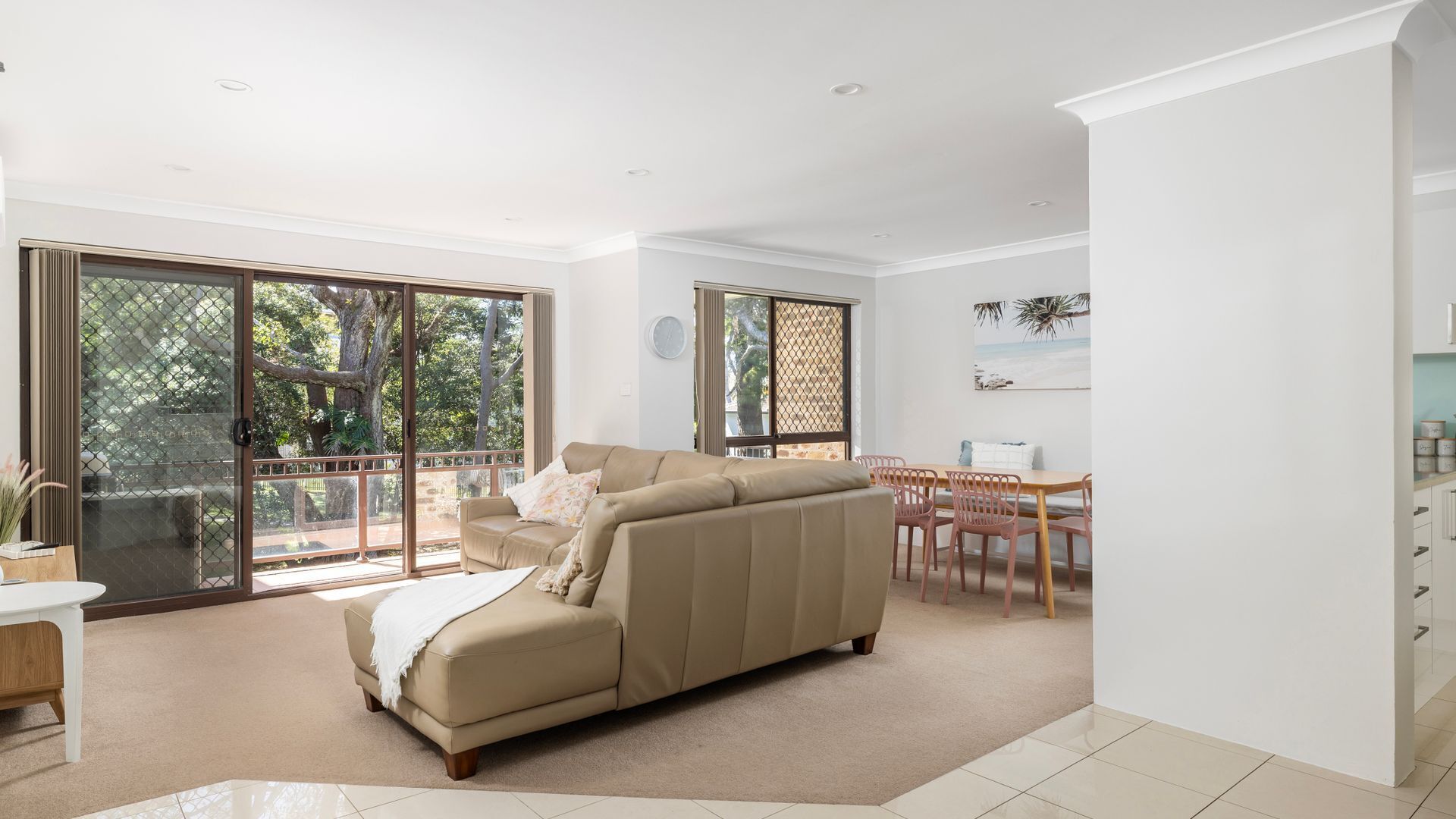 A spacious living room with a beige sofa, a dining area with a wooden table and chairs, and large sliding glass doors leading to an outdoor area.