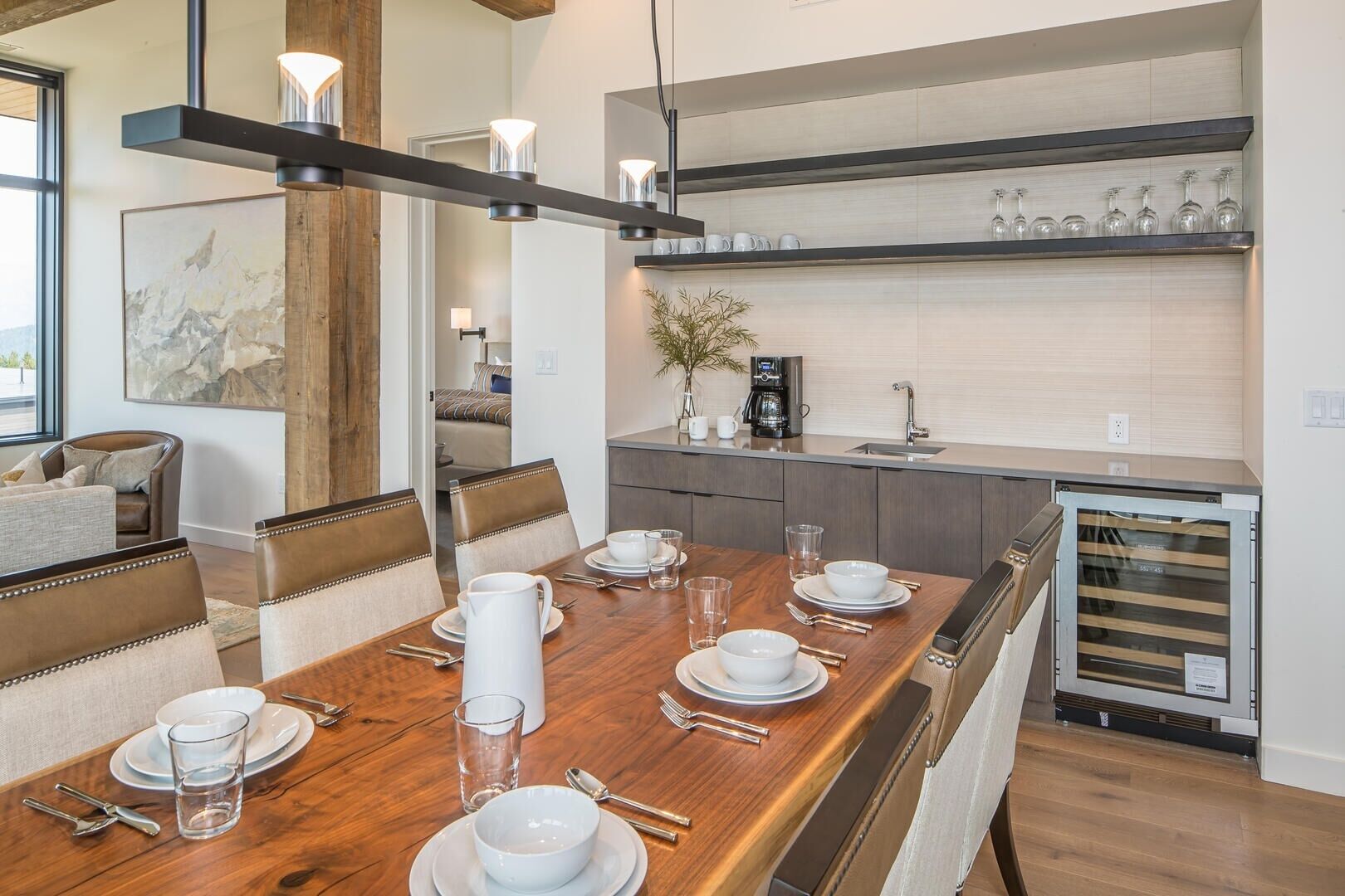 A modern dining room with a wooden table set for four, a coffee machine, and a kitchen area with a sink and shelves above.