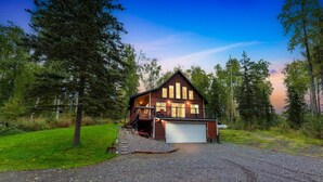 Une cabane en bois à deux étages, dotée d’un garage, entourée d’une allée en gravier et d’une forêt dense.