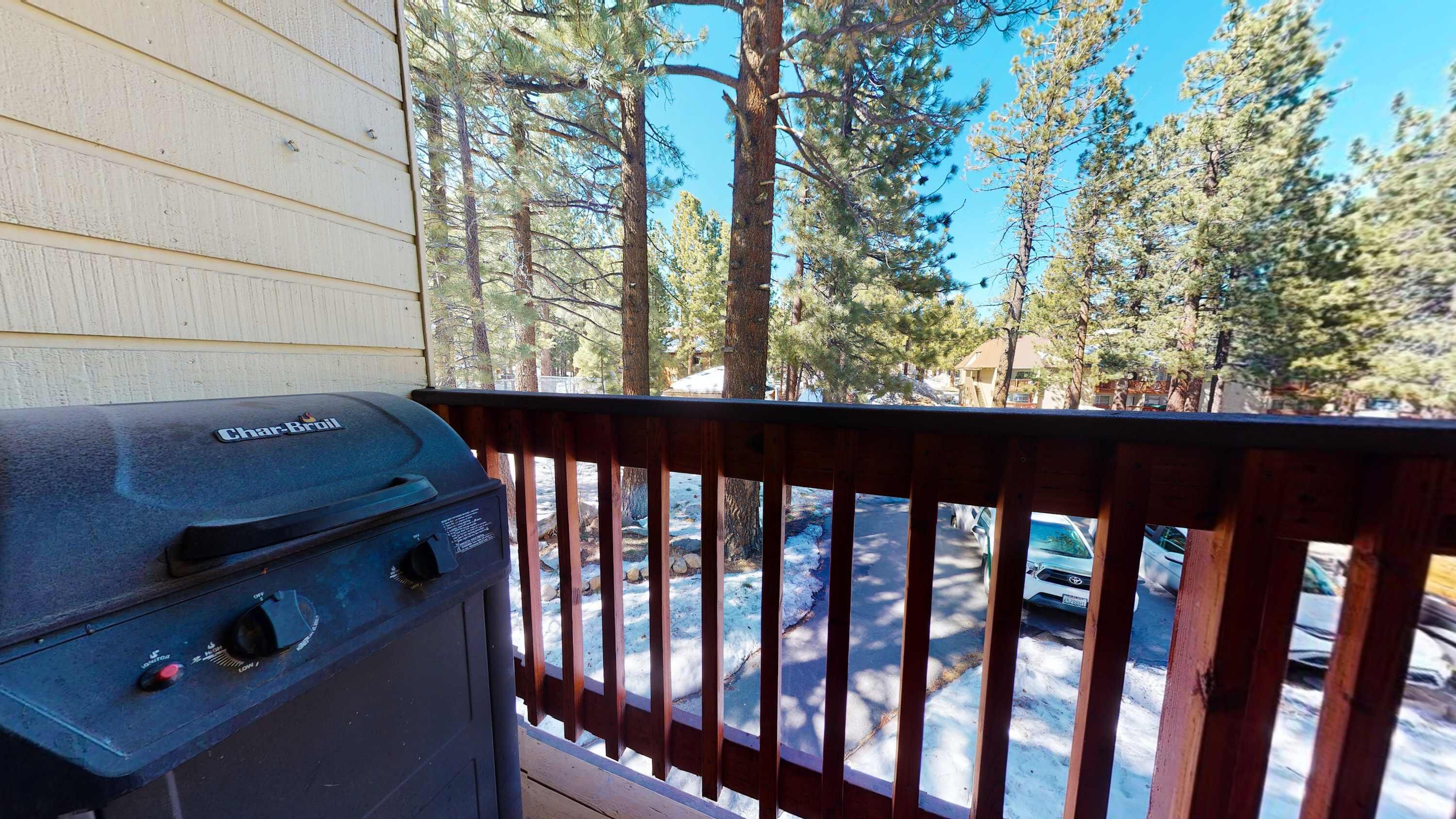 A balcony with a Char-Broil grill, overlooking a street with parked cars and tall pine trees.