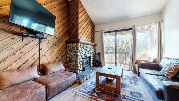 A cozy living room with a stone fireplace, leather sofas, a flat-screen TV, and a large window with a view of trees.