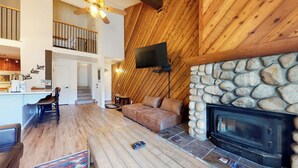 A spacious living area with a stone fireplace, wooden walls, a flat-screen TV, and a staircase leading to an upper level.