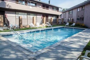 A rectangular swimming pool with a tiled edge, surrounded by a concrete deck. There are white lounge chairs and a small grassy area with a wooden fence. - Pet Friendly | Walk to Shops, Dining & Shuttle (Mammoth Lakes)