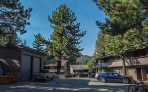 A parking lot with several cars and a motorcycle, surrounded by buildings and tall trees. - Pet Friendly | Walk to Shops, Dining & Shuttle (Mammoth Lakes)
