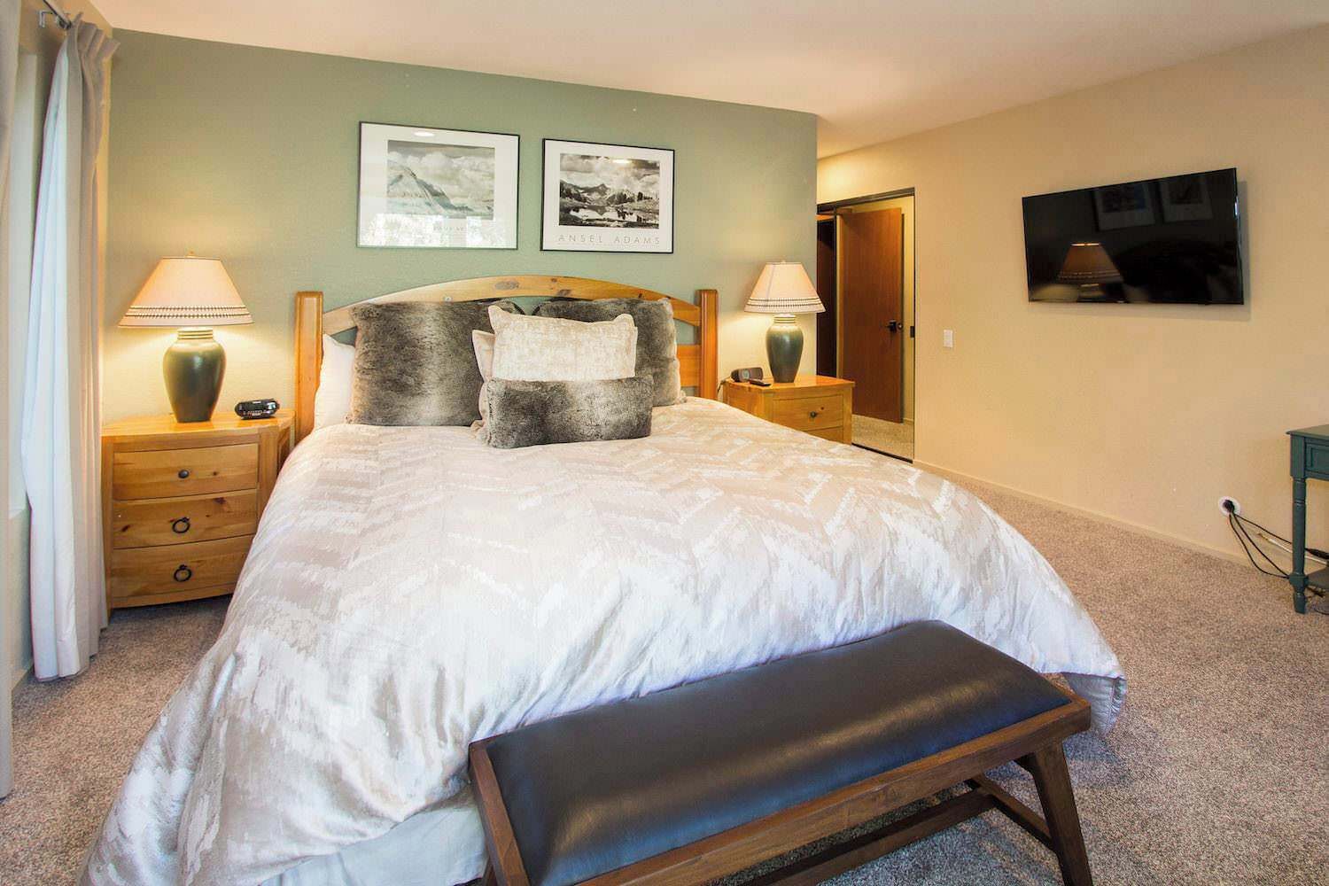 A bedroom with a large bed, two bedside tables, a bench, and artwork on the walls.