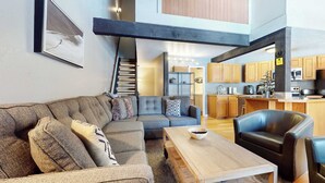 A modern living room with a sectional sofa, a wooden coffee table, and a black leather chair. A staircase leads to an upper level with a kitchen area visible.