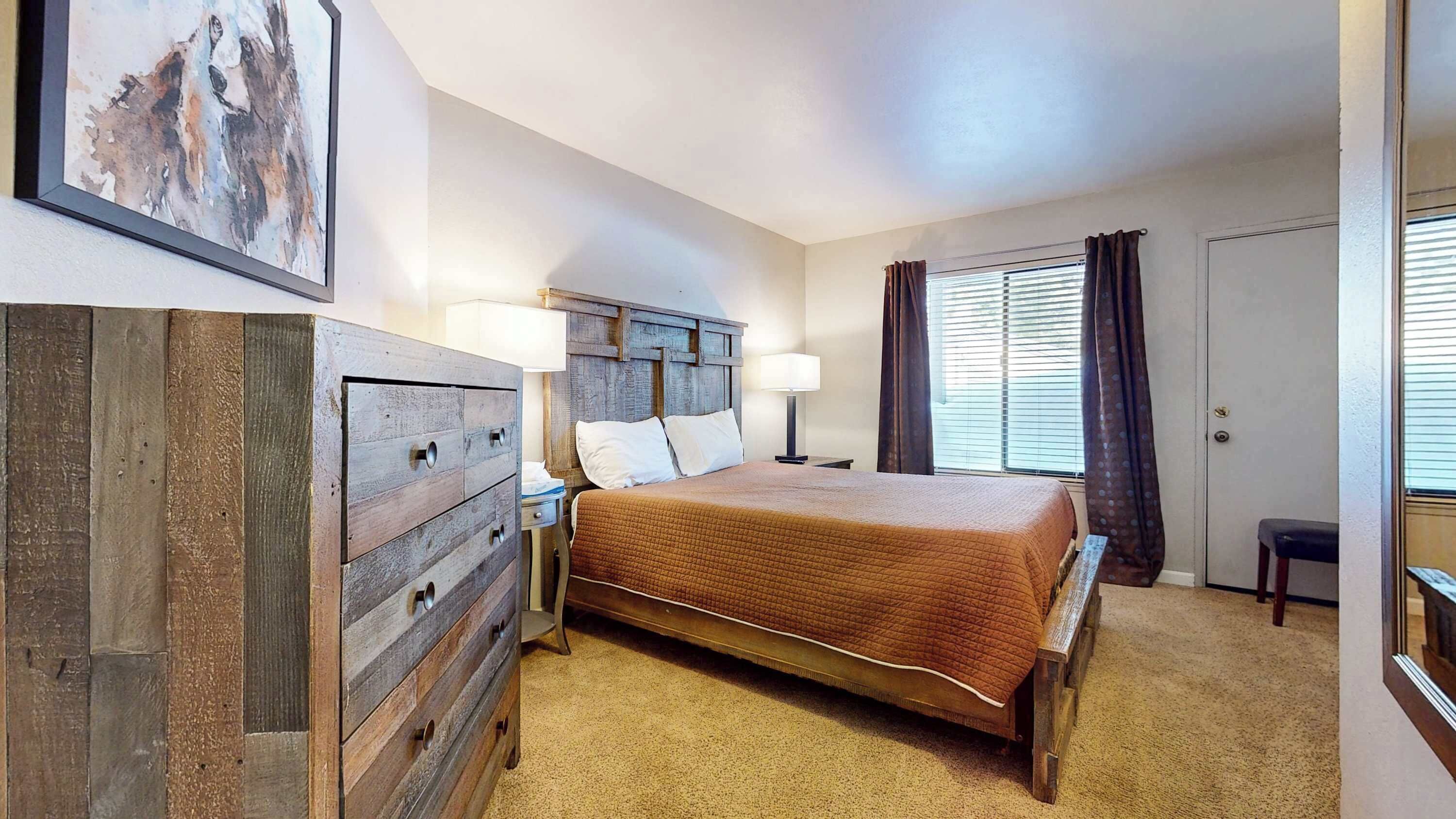 Une chambre à coucher avec une commode en bois, un lit recouvert d’une courtepointe, une table de chevet avec une lampe et une fenêtre avec des rideaux.
