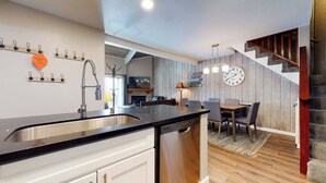 A modern kitchen with a black countertop, stainless steel sink, and a dishwasher. There is a dining area with a wooden table and chairs, and a staircase with wooden steps.