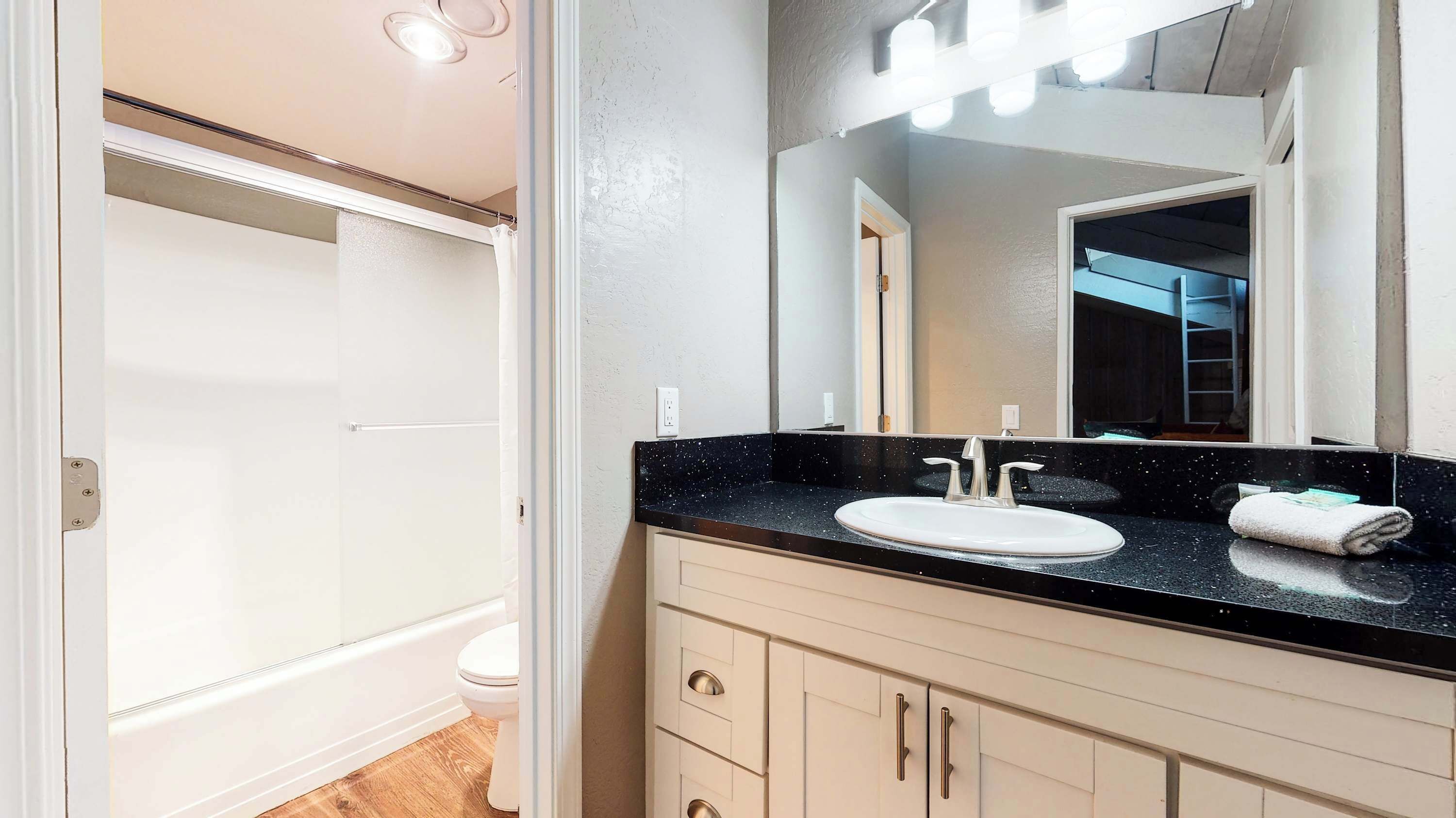 Un baño con encimera negra, lavabo blanco y espejo con luces arriba. Hay una toalla sobre la encimera y un inodoro a la vista.