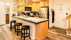 A kitchen with wooden cabinets, a stainless steel refrigerator, a microwave, and a sink. There are three stools and a hanging light fixture.