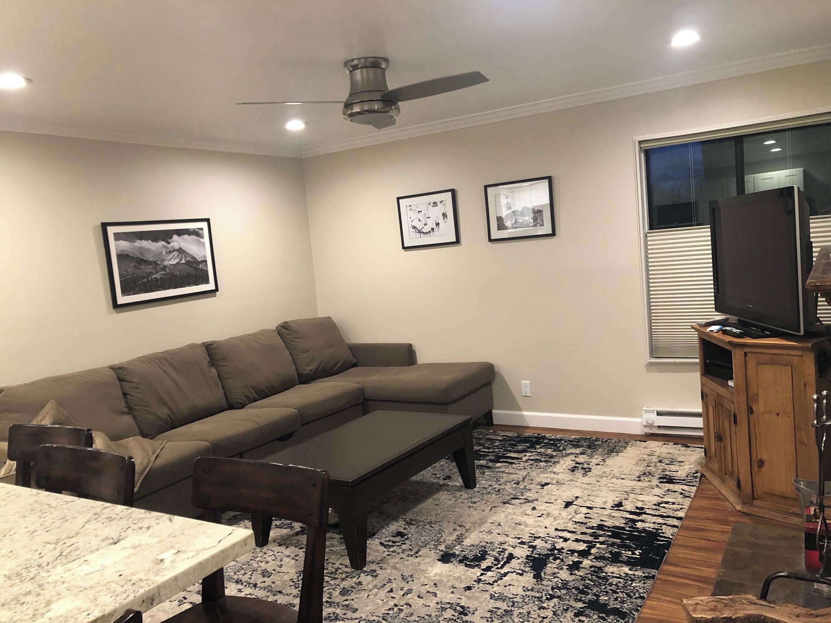 A living room with a sectional sofa, a dining table, a ceiling fan, framed pictures on the wall, and a television on a wooden stand.