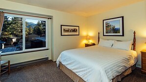 A hotel room with a large bed, two bedside tables with lamps, a window with a view of trees and mountains, and two framed pictures on the wall. - Horizons 4 | Year-Round Outdoor Activities! (Mammoth Lakes)