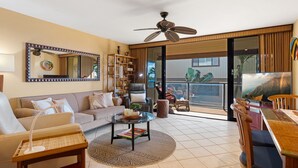 A spacious living room with a sofa, coffee table, and dining area. A large mirror and ceiling fan are visible.