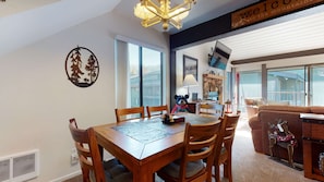 Uma sala de jantar com mesa e cadeiras de madeira, uma estatueta de urso e uma placa com a inscrição 'Bem-vindos às montanhas'.