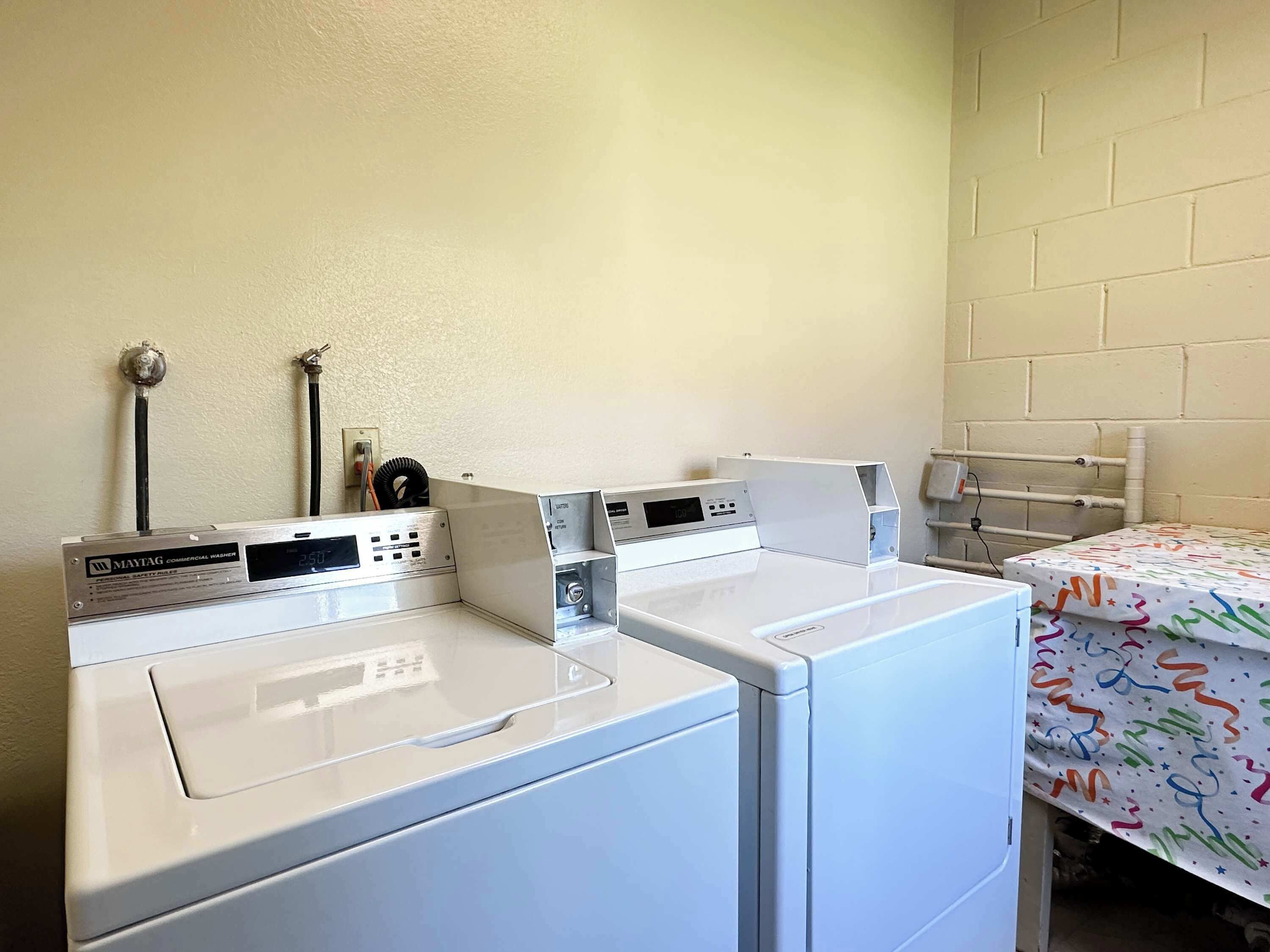 A laundry room with two white front-loading washers and dryers, a white brick wall, and a bed with a colorful bedspread.
