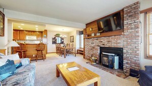 A spacious living room with a fireplace, a dining area with wooden chairs, and a kitchen with wooden cabinets. - Walk to Mammoth Ski Lifts | Ski Storage | Sleeps 6 (Mammoth Lakes)
