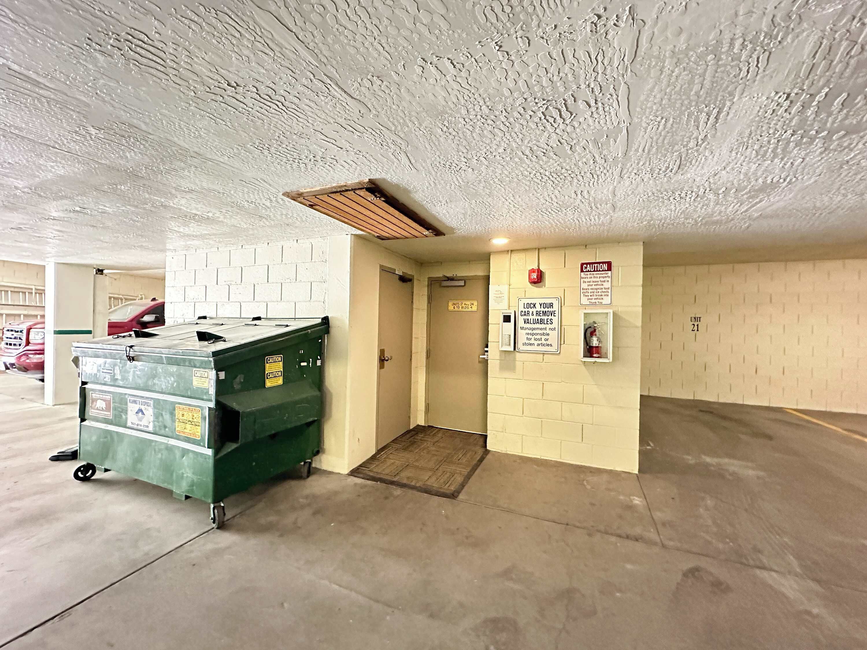 Un parking avec une benne à ordures verte, un extincteur et des panneaux d’avertissement sur les murs.