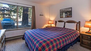 A bedroom with a large window, a plaid bedspread, two bedside lamps, a framed picture on the wall, and a dresser.